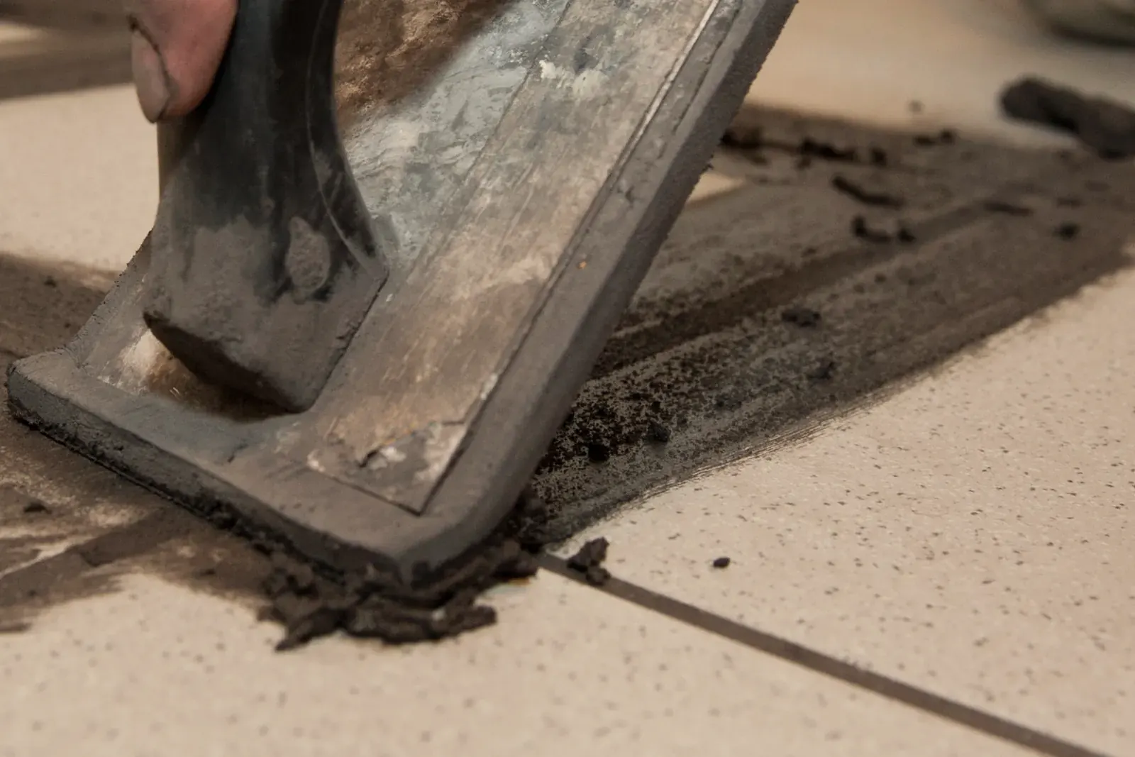 A rubber float pushes dark grout diagonally across the joints of light floor tiles
