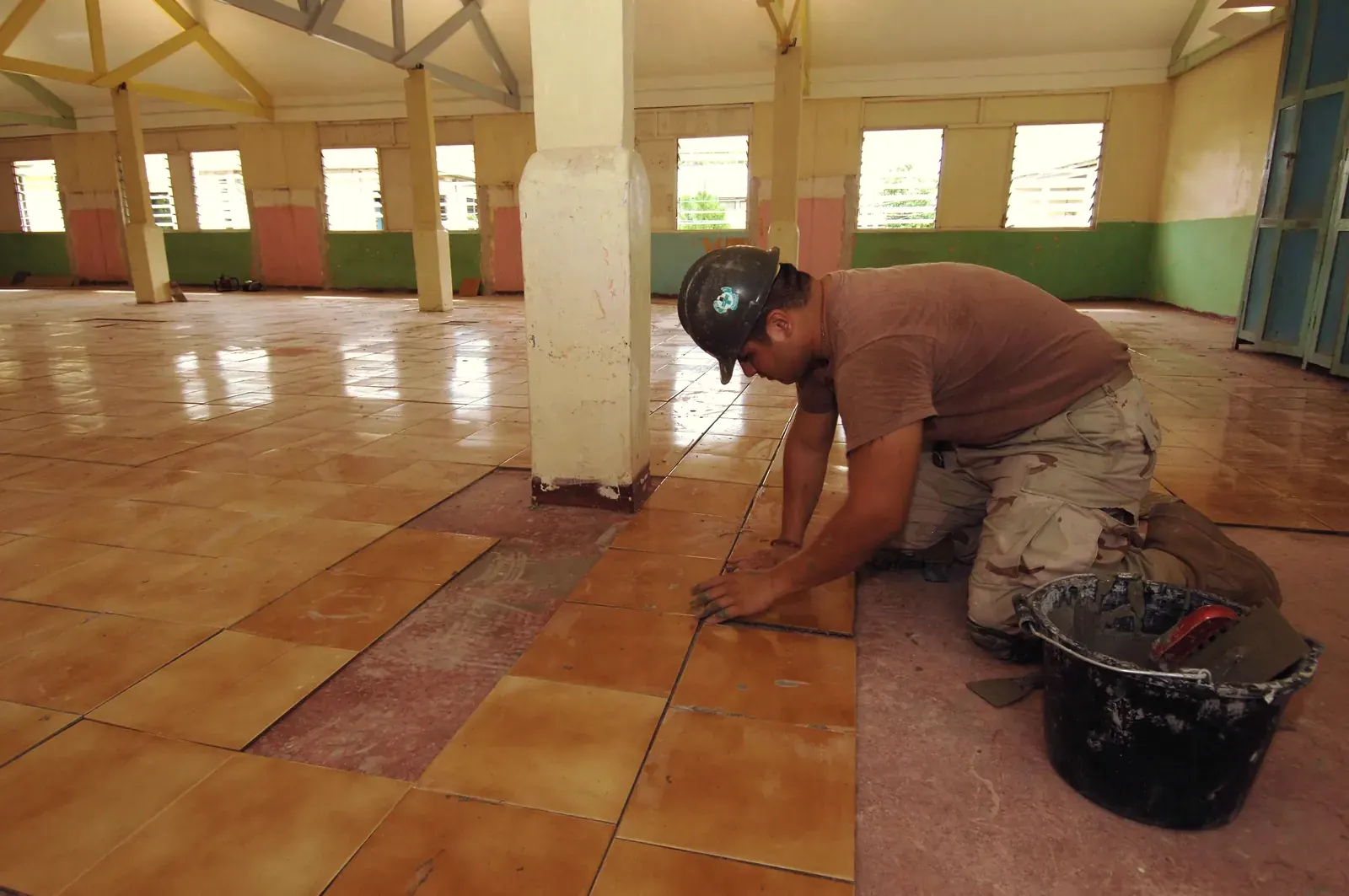 A tiler kneels on the floor of a large hall and sets a ceramic floor tile into the adhesive bed, his bucket beside him