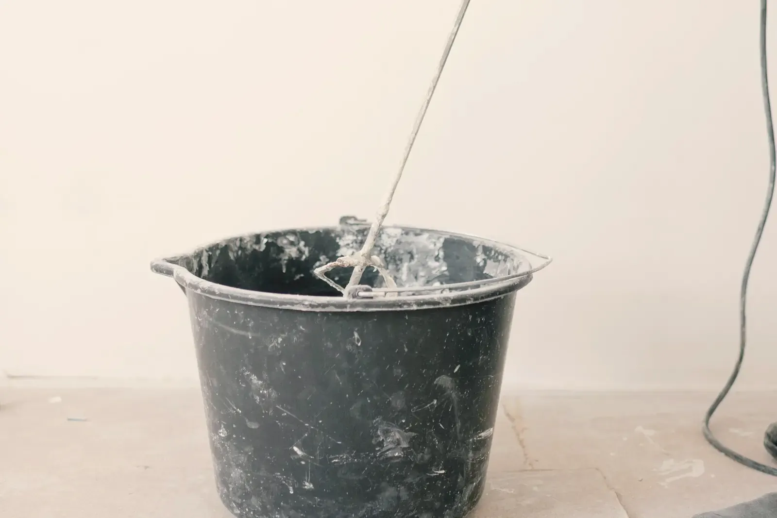 A mixing paddle stands in a mortar bucket in front of a white wall, the drill cable beside it
