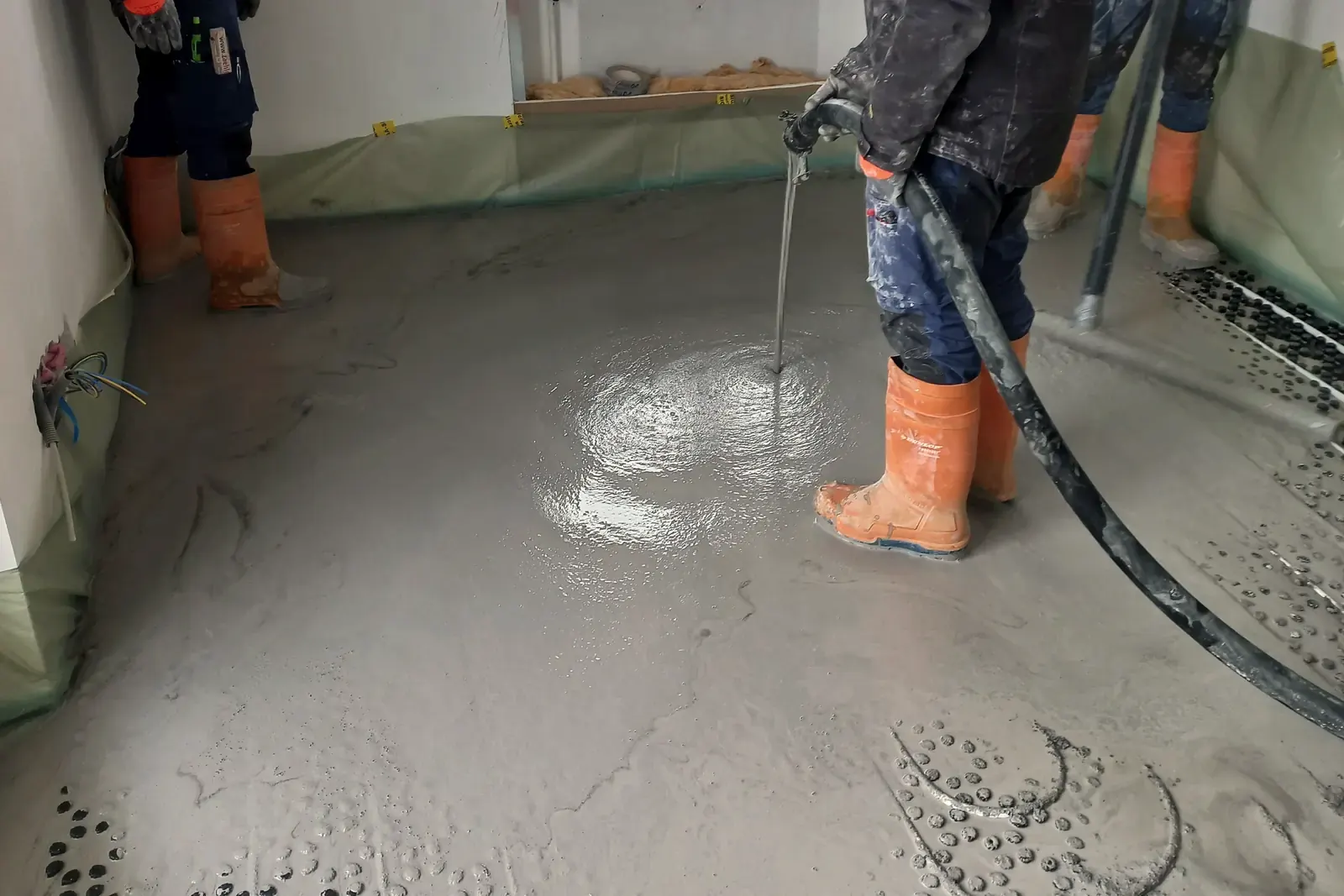Workers in rubber boots pour liquid floor compound from a pump hose in a room lined with edge foil