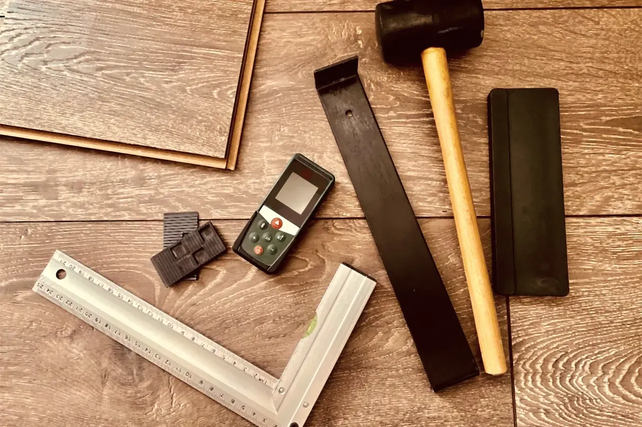 Laminate tools on a wood-look floor: pull bar, rubber mallet, tapping block, wall spacers, laser distance meter and square