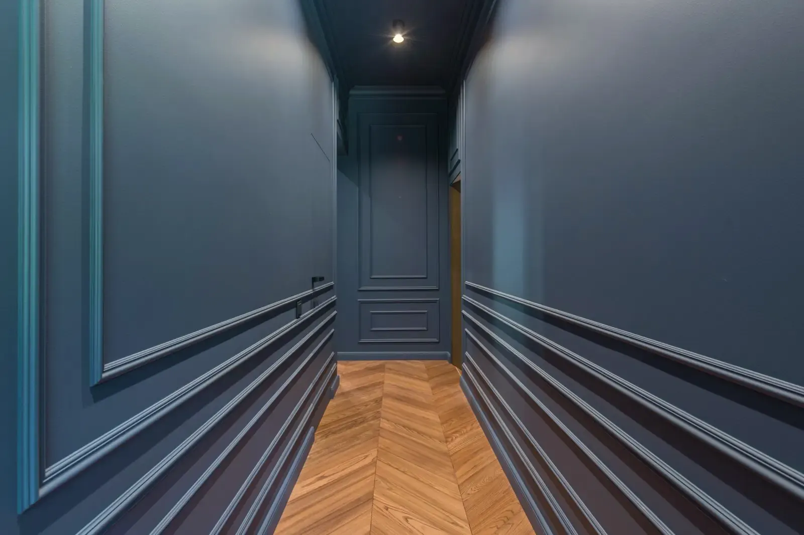 A narrow corridor with blue walls and an oak chevron floor whose points run along the corridor
