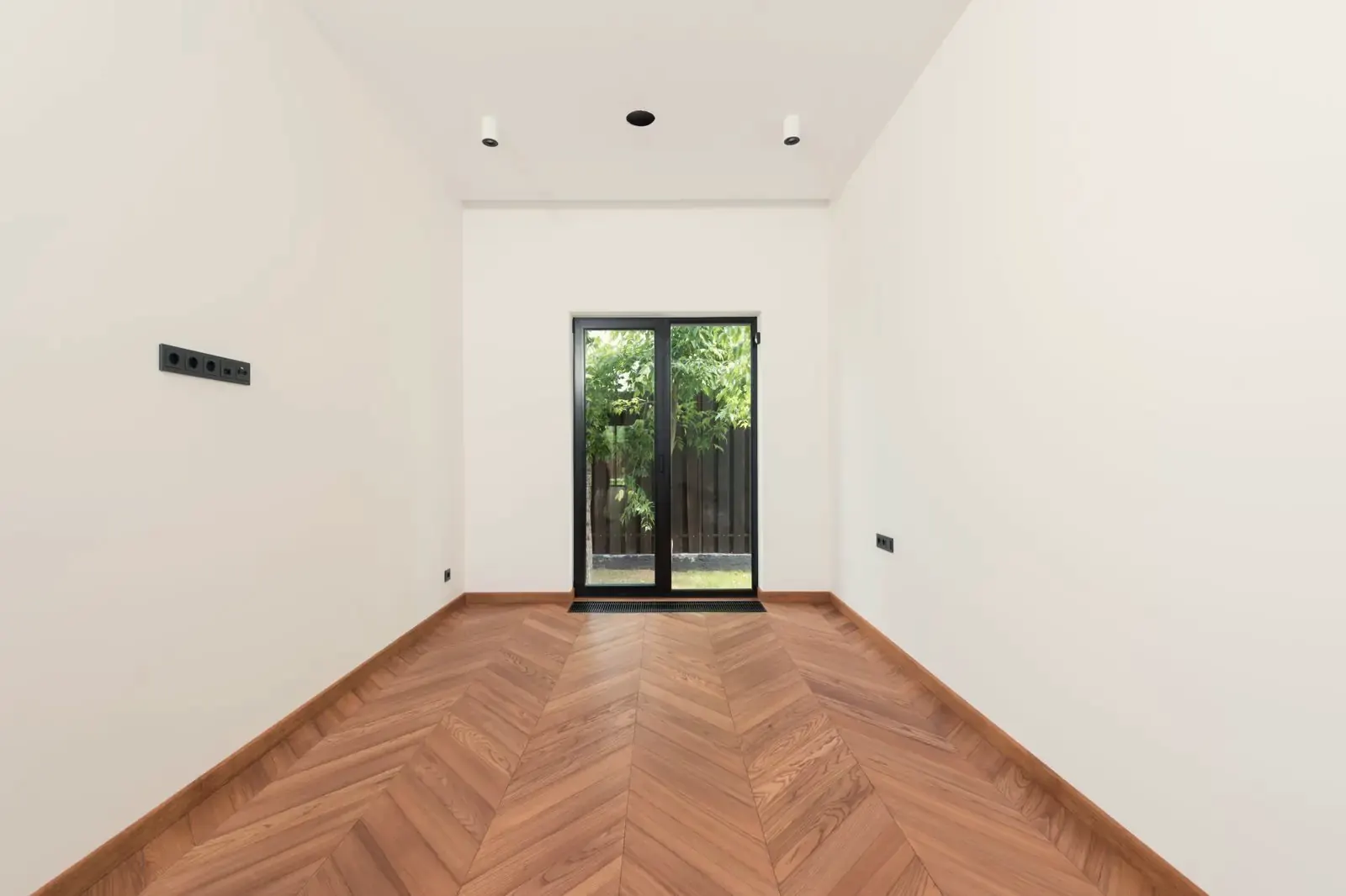 A long, empty room with white walls and a freshly laid chevron floor running towards the glass door at the far end