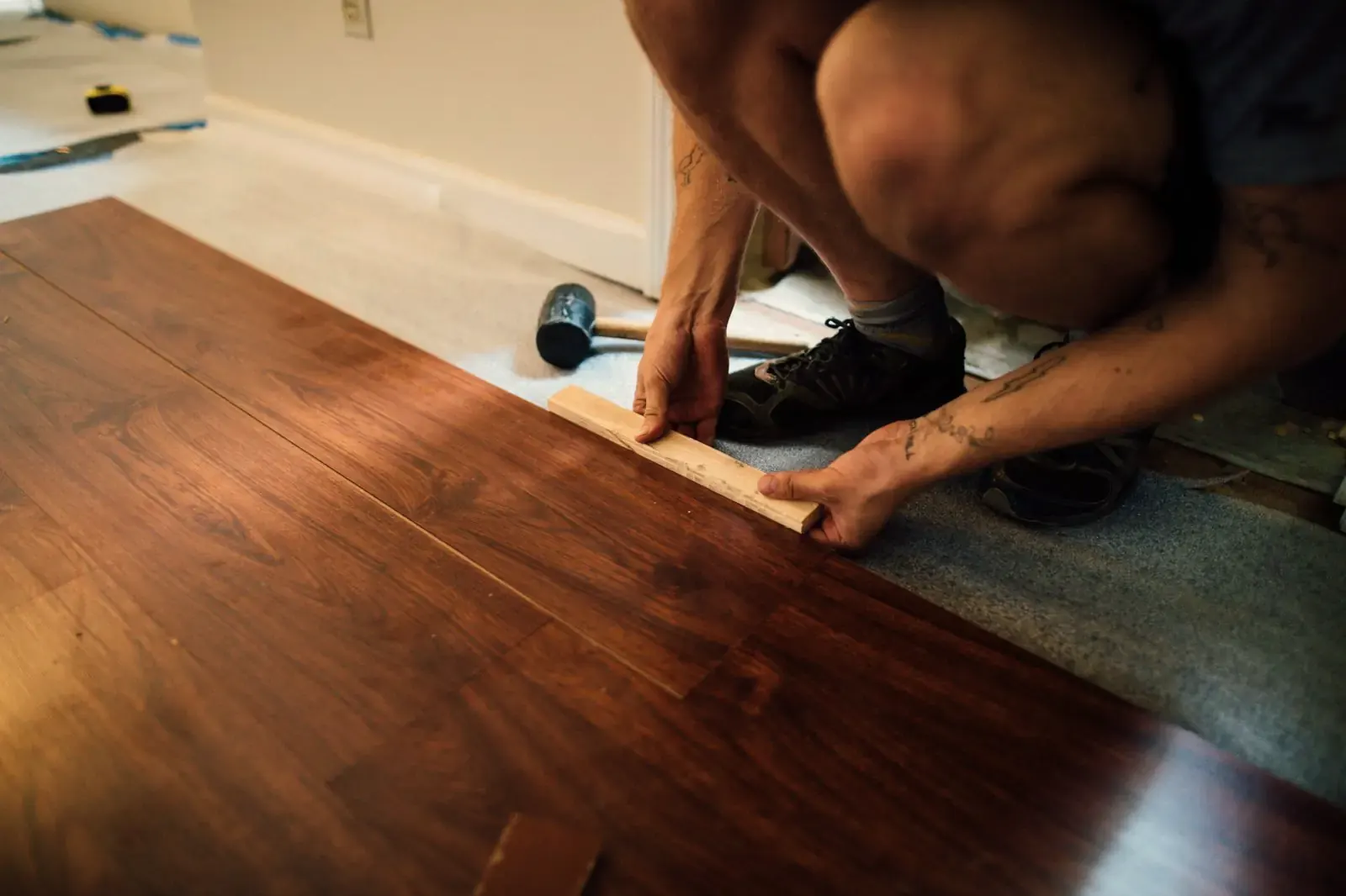 A fitter kneels on a laminate floor and closes a plank row by hand, a rubber mallet beside him and the underlayment rolled out ahead