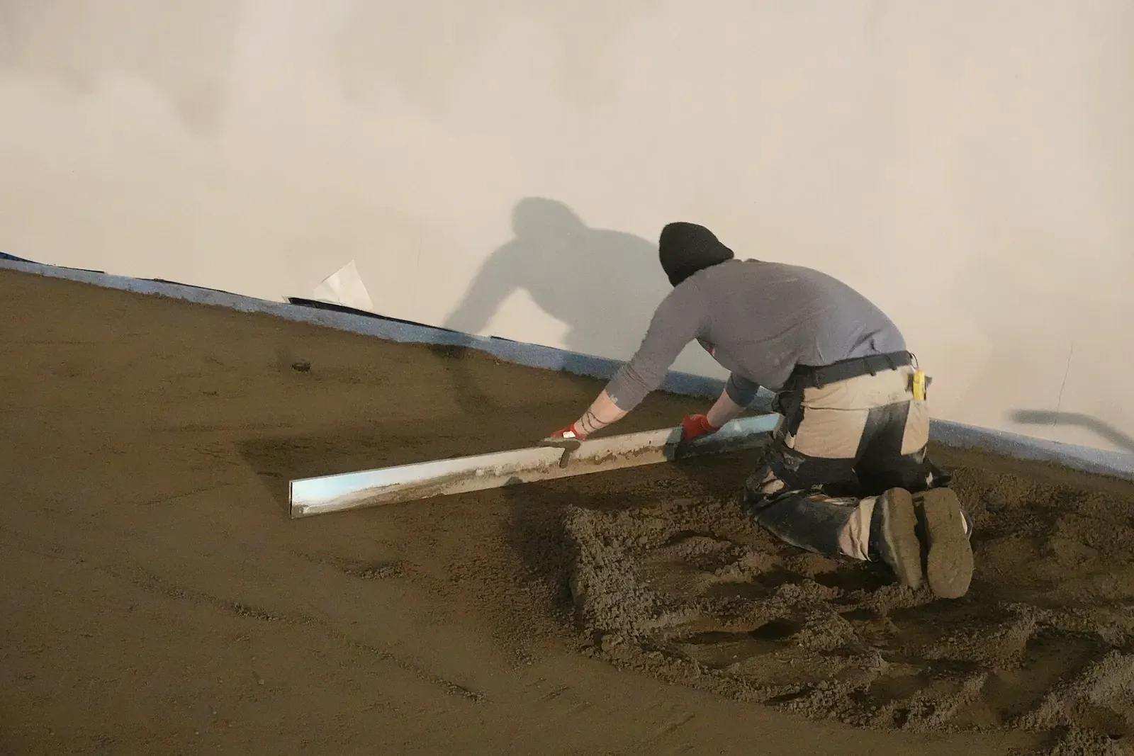 Un operario de rodillas pasa una regla larga sobre una solera de arena y cemento recién echada, junto a una pared blanca con banda perimetral aislante