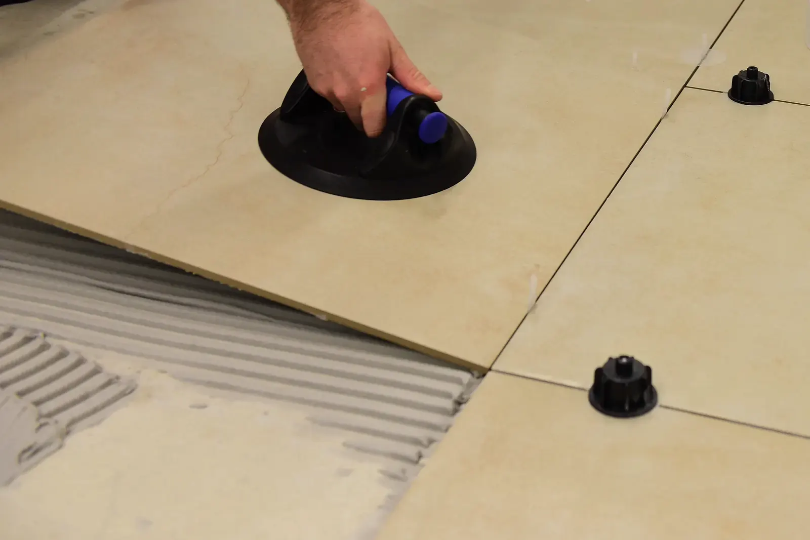 A hand sets a large floor tile with a suction lifter onto combed adhesive, leveling clips in the joints