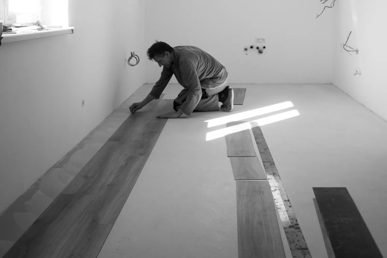 Um instalador ajoelhado junto à parede assenta a primeira fila de tábuas de vinil sobre uma base limpa, com mais tábuas preparadas ao lado, fotografia a preto e branco