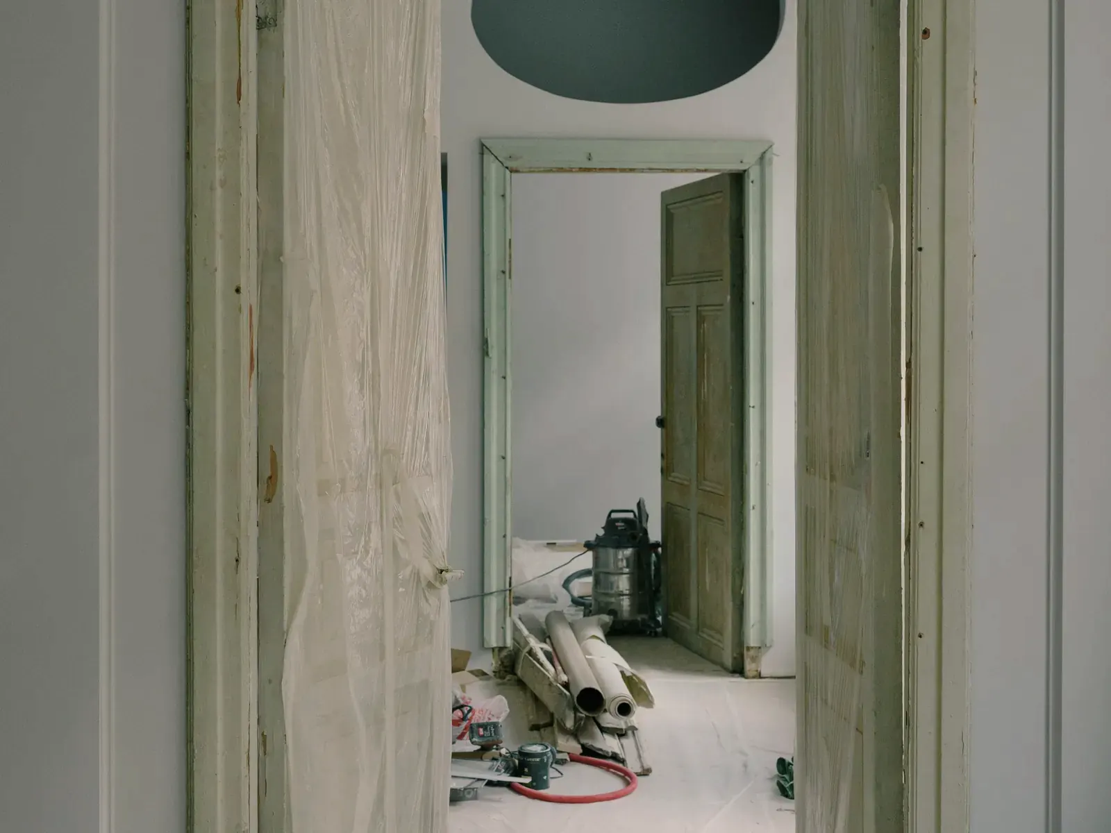 Vista a través de una puerta doble abierta en un piso antiguo, las hojas envueltas en film, el suelo cubierto con manta protectora y al fondo herramientas y un aspirador