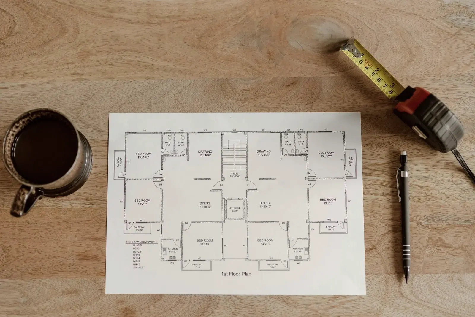 Printed floor plan on a wooden table with a tape measure, a pencil and a cup of coffee