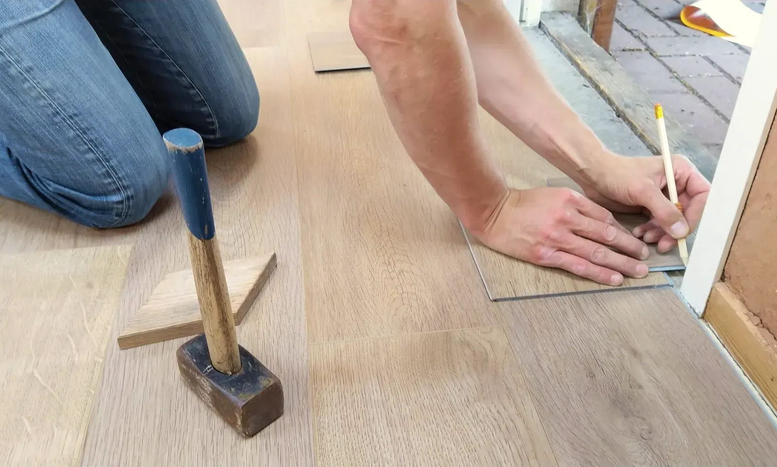 Installer marking a laminate plank for cutting at the door frame, hammer and laid planks in view