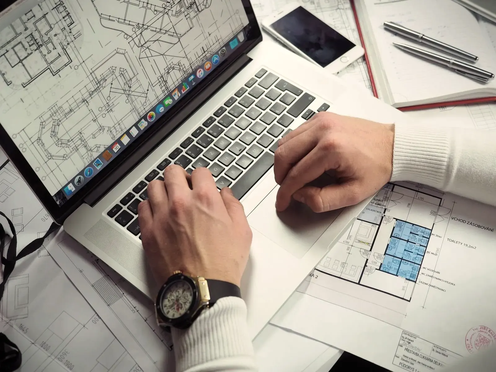 Hands typing on a laptop with a building plan on screen, printed floor plans spread around it