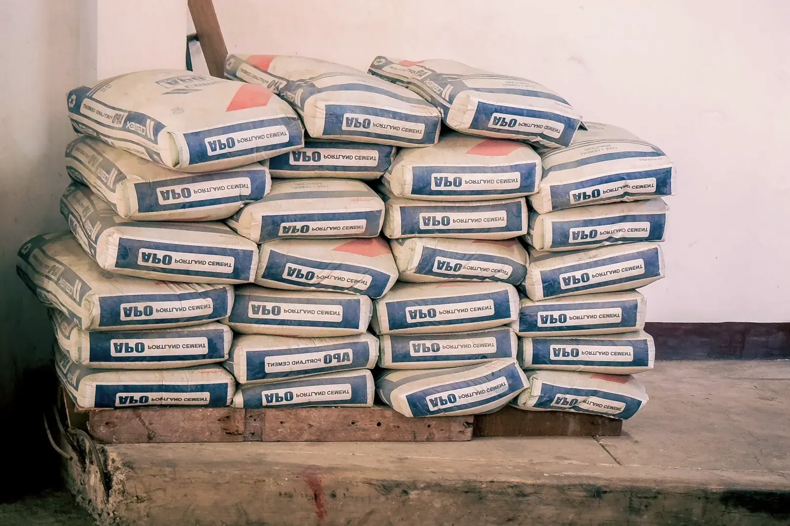 Una pila ordenada de sacos de cemento cerrados sobre un palé de madera junto a una pared blanca en un almacén