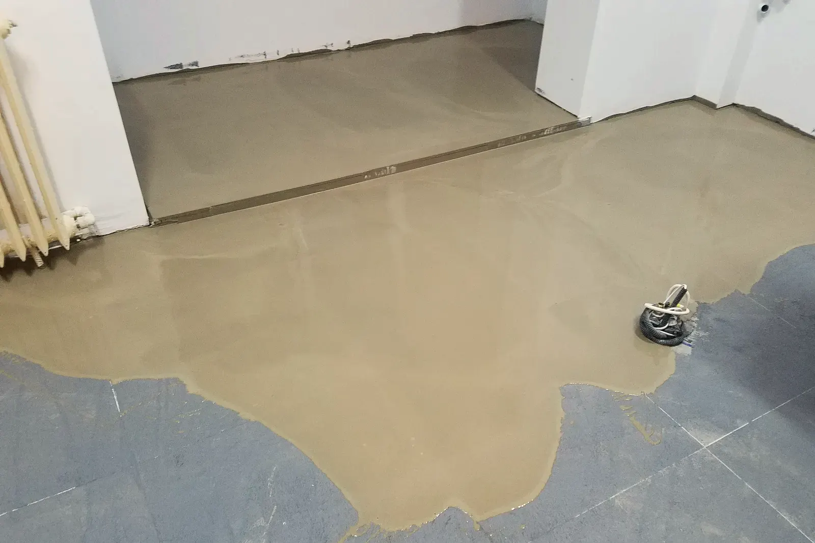 Pasta autonivelante recién vertida se extiende sobre baldosas antiguas en una habitación blanca
