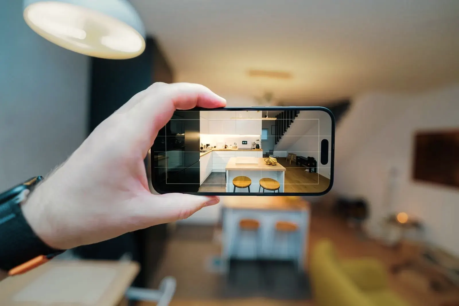 A hand holds a smartphone up to capture a living room and kitchen, the room visible on the screen