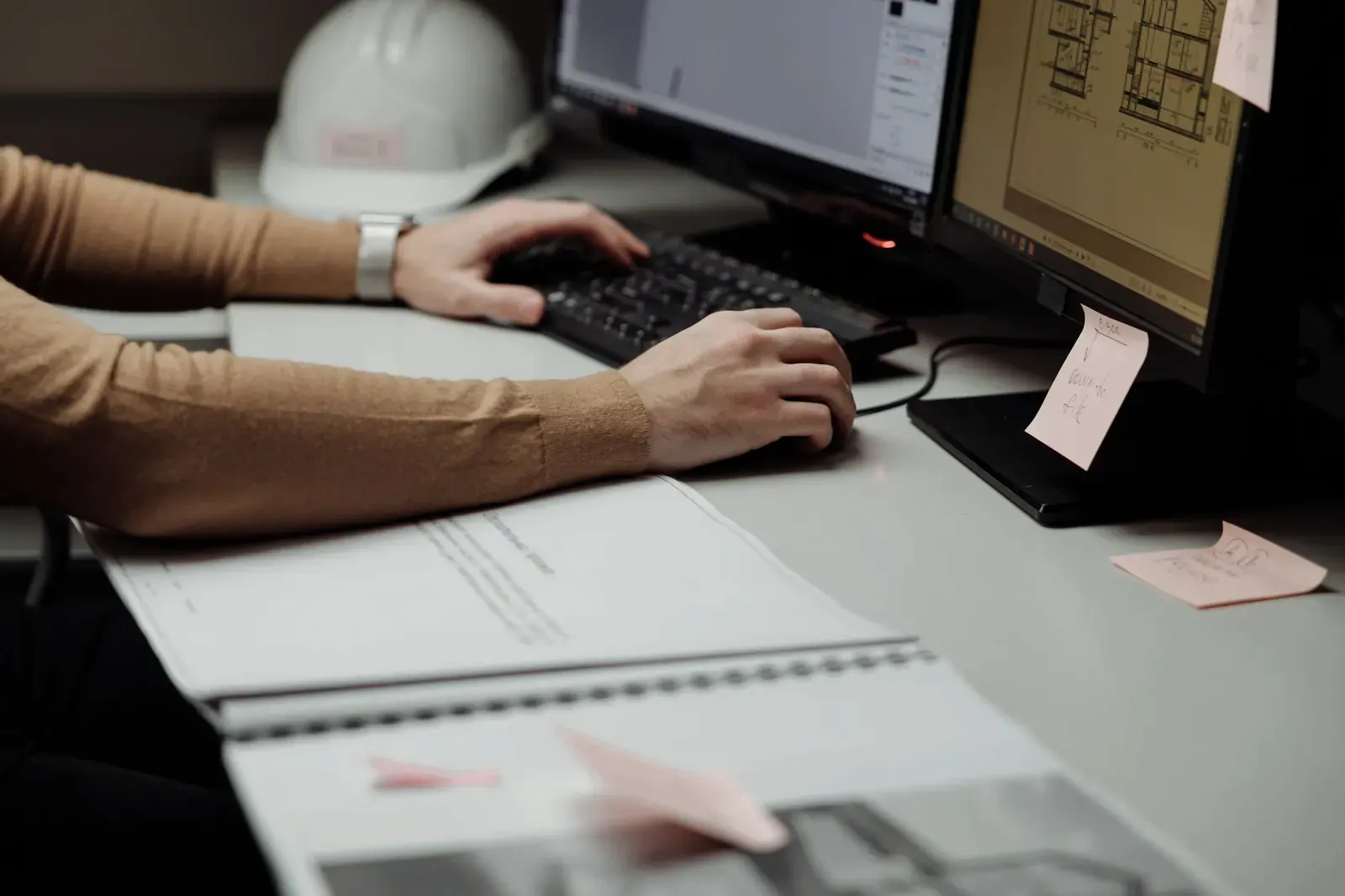 Hands at a keyboard in front of two screens showing building plans, a printed plan set and a white hard hat on the desk