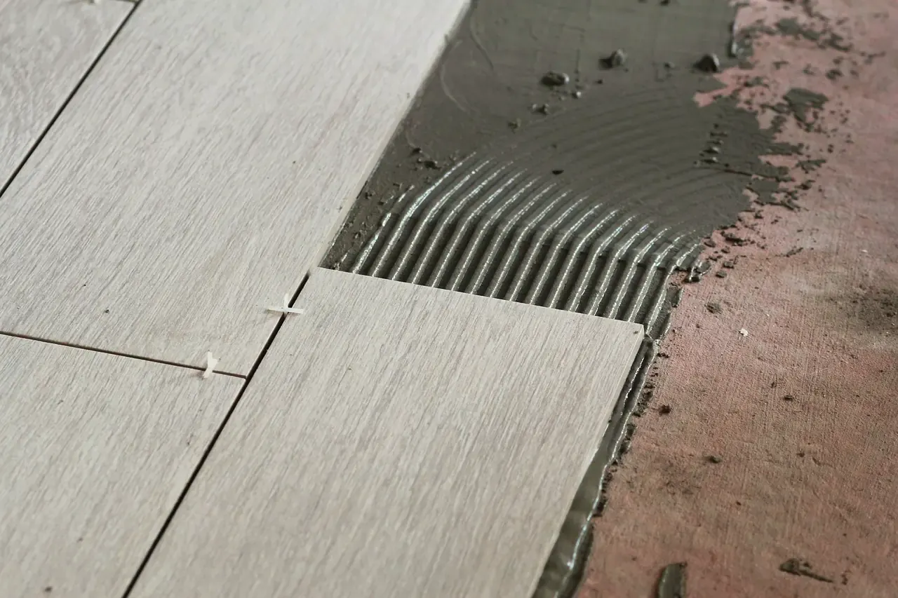 Colla per piastrelle stesa a righe dritte con la spatola dentata su un pavimento interno, accanto a piastrelle effetto legno appena posate con distanziatori