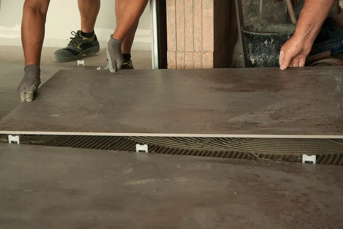 Two tilers set a large-format floor tile onto notched adhesive with levelling clips in a room under renovation
